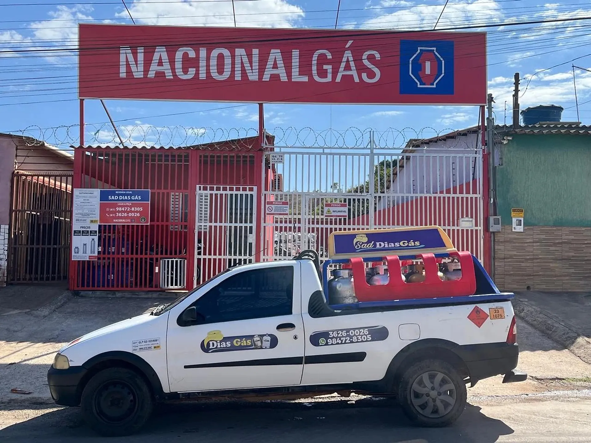 Picape branca da Sad Dias Gás, com o letreiro Sad Dias Gás no teto e a gaiola vermelha de botijões, estacionada diante da revenda com a placa da Nacional Gás